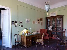 Blick in ein Arbeitszimmer mit Schreibtisch und verglastem Bücherschrank. Hellgrüne Wände, daran verschiedene Zeichnungen und Familienfotos, eine chinesische Deckenlampe.