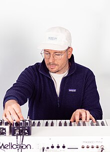 Produzent Shuko in einem weißen Raum, sitzend, an einem weißen Synthesizer