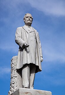 A marble statue of Sir George Grey atop a granite pedestal. Sir George is standing with a scroll in his hand and a Maori carving to the rear.