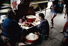 Two women sat at a low table sorting bean sprouts. A large bowl of sprouts is by their side. A child in the background is playfighting, amusing one of the women.