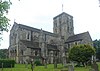 Three-quarter view of a substantial stone church with a square clock tower topped with a weather-vane. The tower has two- and three-light arched openings in each face. A triangular-roofed projection to the right has several round-arched windows. The main body of the church extends to the left and has large buttresses topped with spirelets.