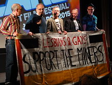 5 white men (4 have glasses) stand holding a banner with handwritten text: "LESBIANS & GAYS SUPPORT THE MINERS". The second man is talking on a microphone. In the background a film screen is partially visible with the text "FILM FESTIVAL [...] 2015".