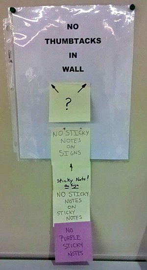 A sign on the wall reading "No thumbtacks on wall", held up with thumbtacks. A sticky note is on the sign with a question mark, and arrows pointing to the thumbtacks. A sticky note is below that sticky note reading "No sticky notes on signs", also on the sign. A sticky note is on that sticky note reading "Sticky note on sign!", with an arrow pointing to the note it is on. A sticky note is on that sticky note reading "No sticky notes on sticky notes". A sticky note is on that sticky note reading "No purple sticky notes", the note is purple.