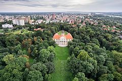 Zdjęcie z lotu ptaka. Pałac Tarnowskich wśród gęstych drzew parku. W tle widoczna szeroka panorama miasta. W tym charakterystyczny, wysoki budynek sądu oraz odległe jezioro i Wisła.