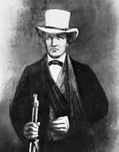 A man in a dark suit with a white hat. His left arm seems to be in a sling. In his right hand he grasps the barrel of a rifle.