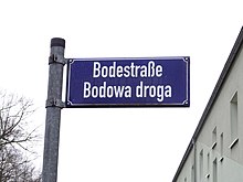 Blaues Straßenschild mit weißer Schrift. Aufschrift: Bodestraße Bodowa droga. In der linken unteren Ecke sind kahle Bäume zu sehen, in der rechten unteren Ecke das Obergeschoss eines Wohngebäudes