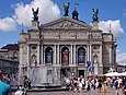 Ein großes helles Gebäude mit Säulen und Skulpturen ist zu sehen. Davor sind viele Menschen und ein Springbrunnen.