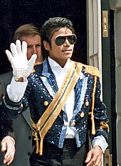 Michael Jackson in a blue suit with a gold diagonal strike, and a white shirt underneath. He has white gloves, waving with his right hand. He is wearing sunglasses.