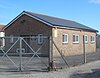 Three-quarter view of a pale brick hut with a shallow tiled roof, standing in asphalt-covered grounds behind a wire fence. There is a recessed entrance door between two windows, and four windows in the longer side.