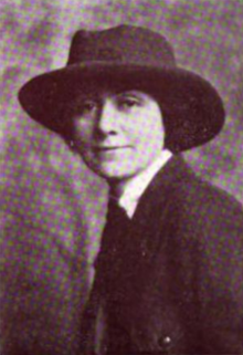 A young white woman wearing a dark brimmed hat and a dark suit with a necktie (may be a Girl Scout uniform, from context)