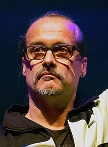 close-up of Alex Christensen wearing a black print t-shirt and black and white Lacoste athletic jacket, looking right of camera with straight face