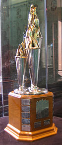 A silver trophy formed of three cones of differing sizes with spiraling "ribbons" at their caps. It has a wooden base with plates engraved with the names of previous winners