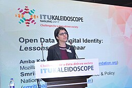 A white woman with short dark brown hair and rectangular glasses stands at a podium with the title of the ITU KALEIDOSCOPE conference on it, she stands in front of a large screen containing the title of her talk