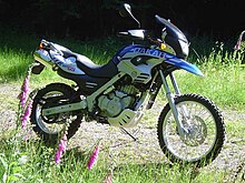 Blue and white BMW F650GS Dakar parked on open ground with tall grass and foxglove flowers in the foreground