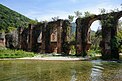 Das Bild zeigt den Ausschnitt einer Brücke des Aquädukts über den Fluss Louros. Sie ist aus Ziegeln gemauert und von ihrer doppelten Bogenreihe stehen noch einige Bögen. Kleine Bäume und Büsche wachsen auf der Brücke und seitlich an den Pfeilern.