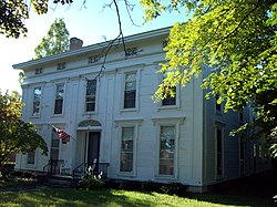 A white house seen from slightly to its right, with a tree branch partially obscuring it. Its front windows and door have some decorative features like columns. An American flag flies from a pole above the main entrance.