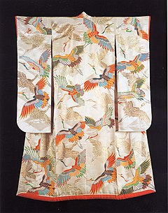 A white satin long-sleeved kimono viewed from the back. The kimono is lined in red silk with a padded skirt and sleeve hem, and is woven with a large, figured pattern of cranes in flight, and is decorated with a dyed and gold foil pattern of cranes in flight of the same size, depicted in a number of colours.