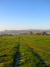 A lightly rutted field in winter, with several buildings in the middle distance and hills behind. The buildings are partly obscured by fences, and consist of farm outbuildings, a large farmhouse with a red-tiled roof and chimneys, a smaller adjacent building in a similar style, and a two-storey house.