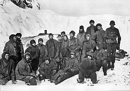 A group of men sitting closely packed together, in heavy winter clothes and wearing hats. Snow and ice on the ground and in the background