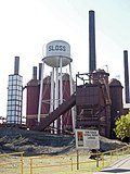 Photograph of the Sloss Blast Furnaces, dominated by the smokestacks and a water tower marked "Sloss". In the foreground is an informational sign discussing the Furnaces' National Historic Landmark status.