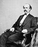 Black and white photo of a balding man with a moustache sitting in a chair. He wears a dark suit of civilian clothes.