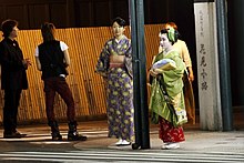 Two young women in kimono stood at a street corner at night. The taller woman wears a casual blue and yellow kimono; the smaller woman is dressed as an apprentice geisha, in a green kimono with green hair accessories and a red underkimono.