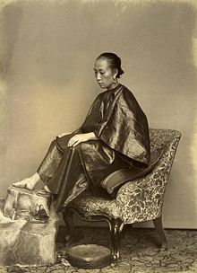 An albumen silver print photograph of a young woman with bound feet; she sits on a chair facing left, her feet - one with a lotus shoe, the other bare - propped up on a stool.