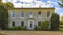 A Grade 2 Listed Georgian country house. Rectangular in shape, with a door in the centre, two windows on either side, and five along the upper floor. Shrubs grow by the corners of the lower floor.