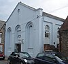 Three-quarter view of a pale blue building with a plain exterior and a large pediment, on a crowded site between two flint structures. The façade has three tall, triple-recessed blank arches, the centre of which is shortened to accommodate an entrance door. A round opening sits just below the pediment. The side has round-arched windows.