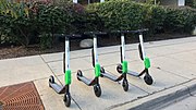 Four electric stand-up scooters lined up on a sidewalk ready for rental. These have black handlebars, white stems, a green battery pack, and gray decks.