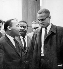 Malcolm X and Martin Luther King Jr. speak to each other thoughtfully as others look on.