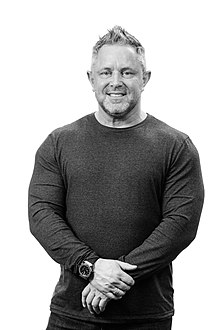 Greyscale image of a Caucasian man with a dark shirt, light, somewhat spiked-upwards hair, and his hands together