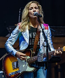 Sheryl Crow standing in front of a microphone and singing while playing guitar