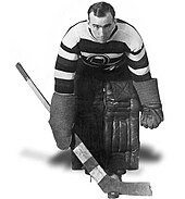 Black and white image of a hockey goaltender holding a stick, wearing pads and a horizontal=striped jersey