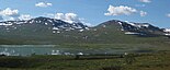 Ein kleiner See inmitten einer spärlich bewachsenen, teils mit Schnee bedeckten Hügellandschaft unter blauem Himmel mit wenigen Wolken