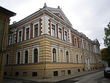 Seitliche Farbfotografie eines zweigeschossigen Gebäudes im Barockstil. Das Erdgeschoss besteht aus gelber Fassade und das Obergeschoss aus brauner Fassade. An allen Fenstern und am Eingang sind weiße Umrandungen. An beiden Dachenden sind Giebelfelder mit Ornamenten.