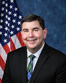 Official House portrait of Mannion smiling in front of the U.S. flag, wearing a black suit, white shirt, and green and blue plaid tie.