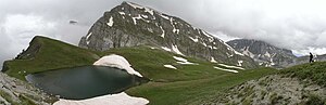 A small lake surrounded by alpine pasture with traces of snow and two mountain peaks in the background: center and right.