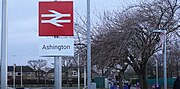 A sign showing a National Rail logo with Ashington written below it with some trees in the background