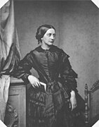 Clara Schumann ist stehend auf einer schwarz-weiß Fotografie abgebildet. Sie lehnt an einem Möbelstück und hält ein Buch in der Hand. Sie trägt ein dunkles Kleid.