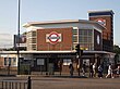 A red-bricked building with a blue sign reading "BOUNDS GREEN" in white letters and several people walking in the foreground all under a blue sky