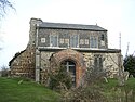 A stone church with a brick porch seen from the south. To the left are the remains of the collapsed tower. The clerestory contains three windows and six carved panels.