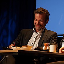 Foto eines an einem Tisch auf einer Bühne sitzenden Mannes