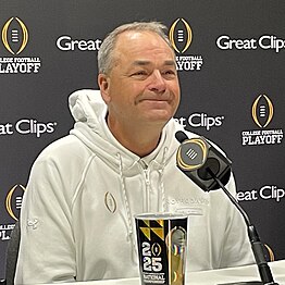 A football coach smiles in front of a microphone.