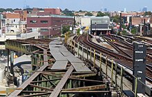 Viel Stahl und Stadt im Blickfeld: In der linken Bildhälfte das teils rostige, teils mit grünem Schutzanstrich versehene Tragwerk einer ehemaligen Hochbahnstrecke. Es basiert auf einem im Querschnitt rechteckigen Körper aus vier Trägern, die durch diagonale Verstrebungen gegenseitig zu einem stabilen Gesamttragwerk verbunden sind. Auf ihm lagen direkt die Schwellen und darauf die Schienen der Hochbahnstrecke. Man sieht hier ein solches Tragwerk nach links in der Bildmitte verlaufen und dort mechanisch abgeschnitten enden. Die rechts Bildhälfte wird von einem Rampenbauwerk mit vier Gleisen bestimmt, entlang dem die Betrachterin abwärts auf die nebeneinander angeordneten Tunnelportale der Culver Line blickt. An den Seiten der Rampe sind Straßen, Laternen, Schilder und Gebäude zu sehen, in der Ferne einzelne Hochhäuser vor diesigem Himmel.