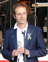 Luigi Gaggero mit einem Mikrofon, am Jackett eine Schleife in den ukrainischen Nationalfarben.