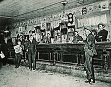 Multiple men in suits standing in front of a bar with no stools and multiple bartenders. The back of the bar has wallpaper, mirrors and posters.