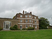 A three storey brown brick building with a cupola, and a single storey extension on the left, the foreground is a green lawn