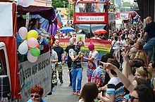 Three people in ski masks march in a LGBT Pride parade. One is holding a sign with a religious-style icon portrait of a member of the group.