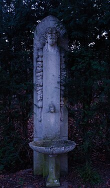 Ein Trinkbrunnen im öffentlichen Raum, vor ein paar Bäumen. Oben am Trinkbrunnen ist eine abbaend hergestellte Brunnenfigur, die eine Bildnisplastik ist.